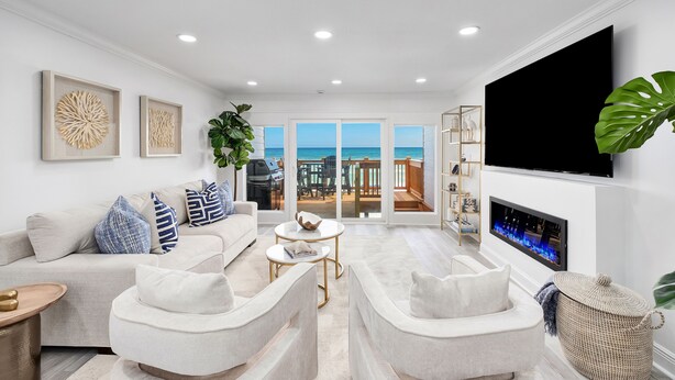 Un vaste salon comprenant un canapé blanc, deux fauteuils, une table basse et un téléviseur à écran plat fixé au mur. Une grande fenêtre offre une vue sur l’océan.