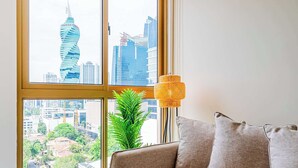 A room with a large window offering a view of a city skyline, a woven lamp, and a sofa with cushions. - w* | Charming 1BR in the Heart of Obarrio (Panamá)