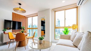 A modern living room with a sofa, a dining area with chairs, a TV mounted on the wall, and large windows offering a city view. - w* | Charming 1BR in the Heart of Obarrio (Panamá)