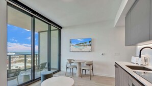 Een moderne keuken met een groot eiland, een eethoek met een ronde tafel en stoelen, en een grote schuifpui die leidt naar een balkon met uitzicht op de stad en de oceaan.