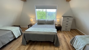 Une chambre à coucher avec un grand lit, deux tables de chevet avec des lampes, une commode et une fenêtre donnant sur des arbres.