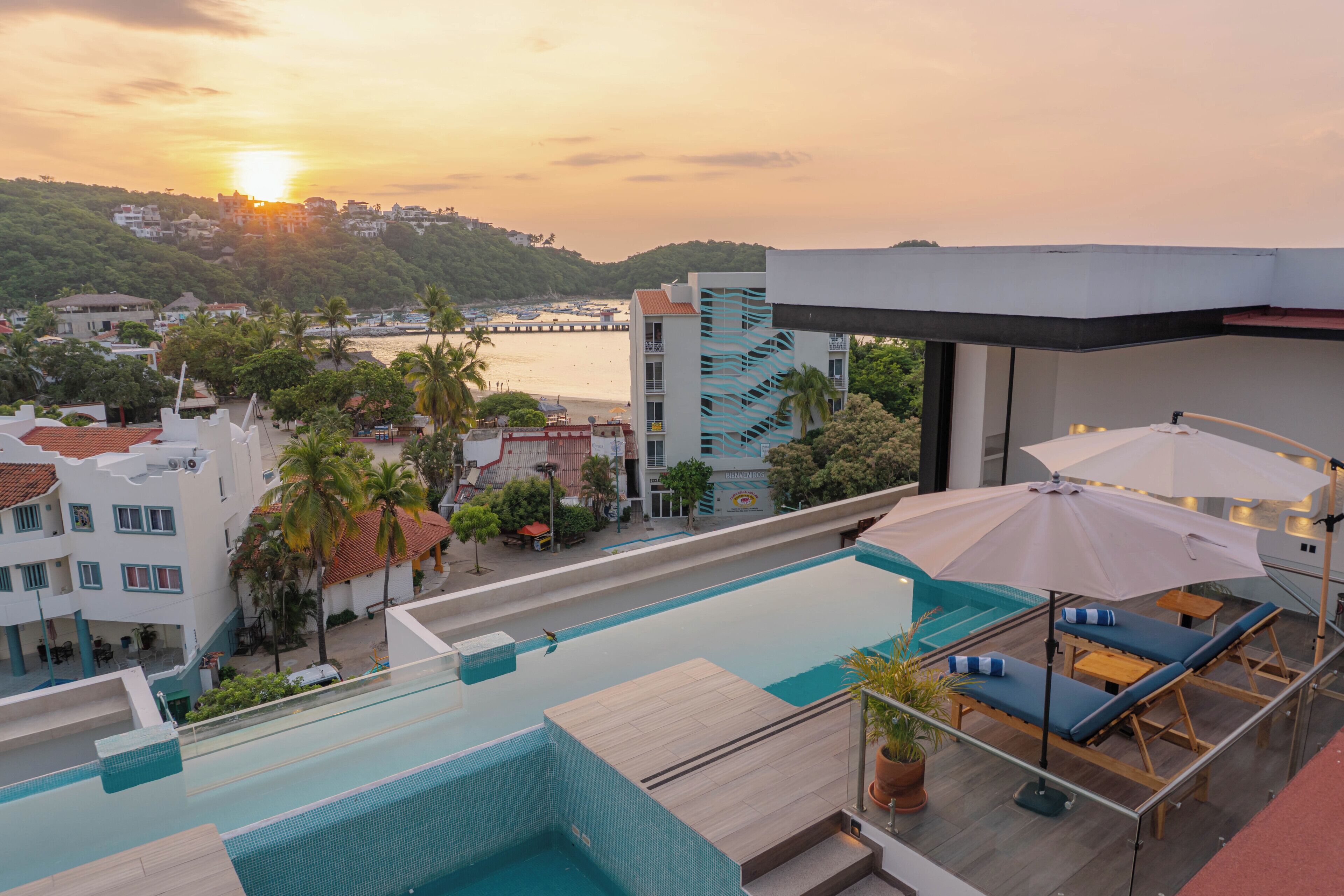 A rooftop pool with lounge chairs and umbrellas, overlooking a coastal area with buildings and a sunset.