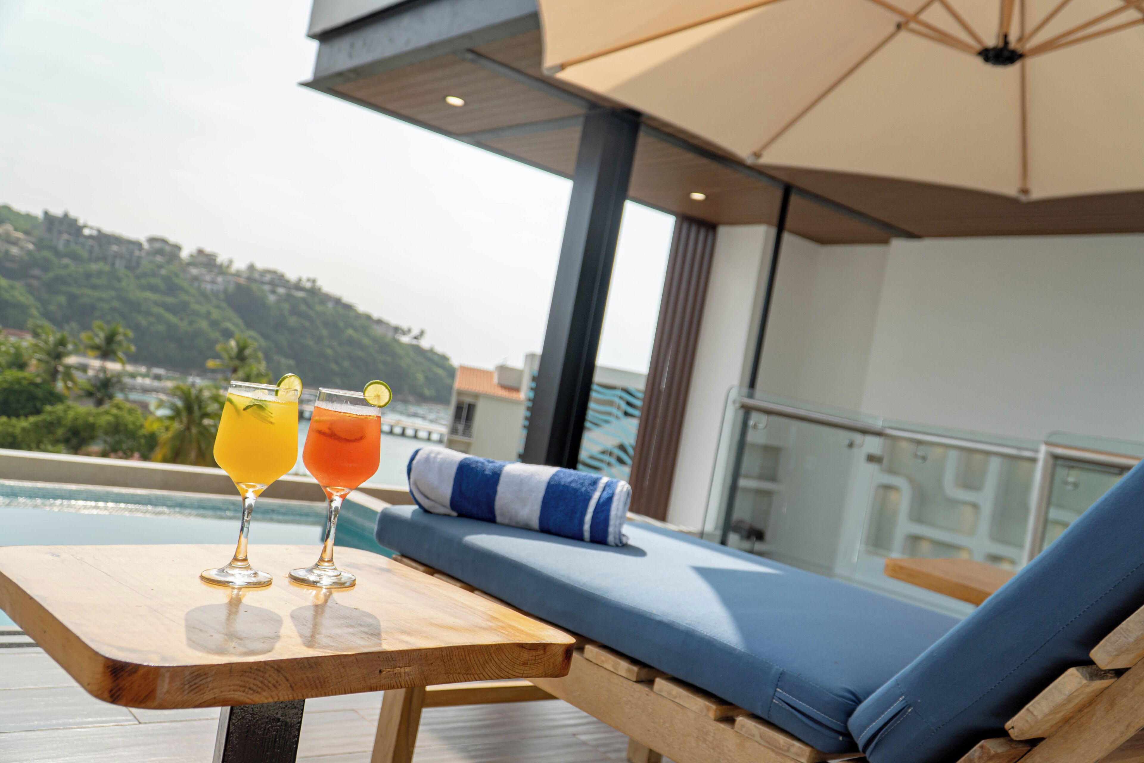 A wooden table with two drinks and a blue cushioned lounge chair on a balcony overlooking a pool and greenery.