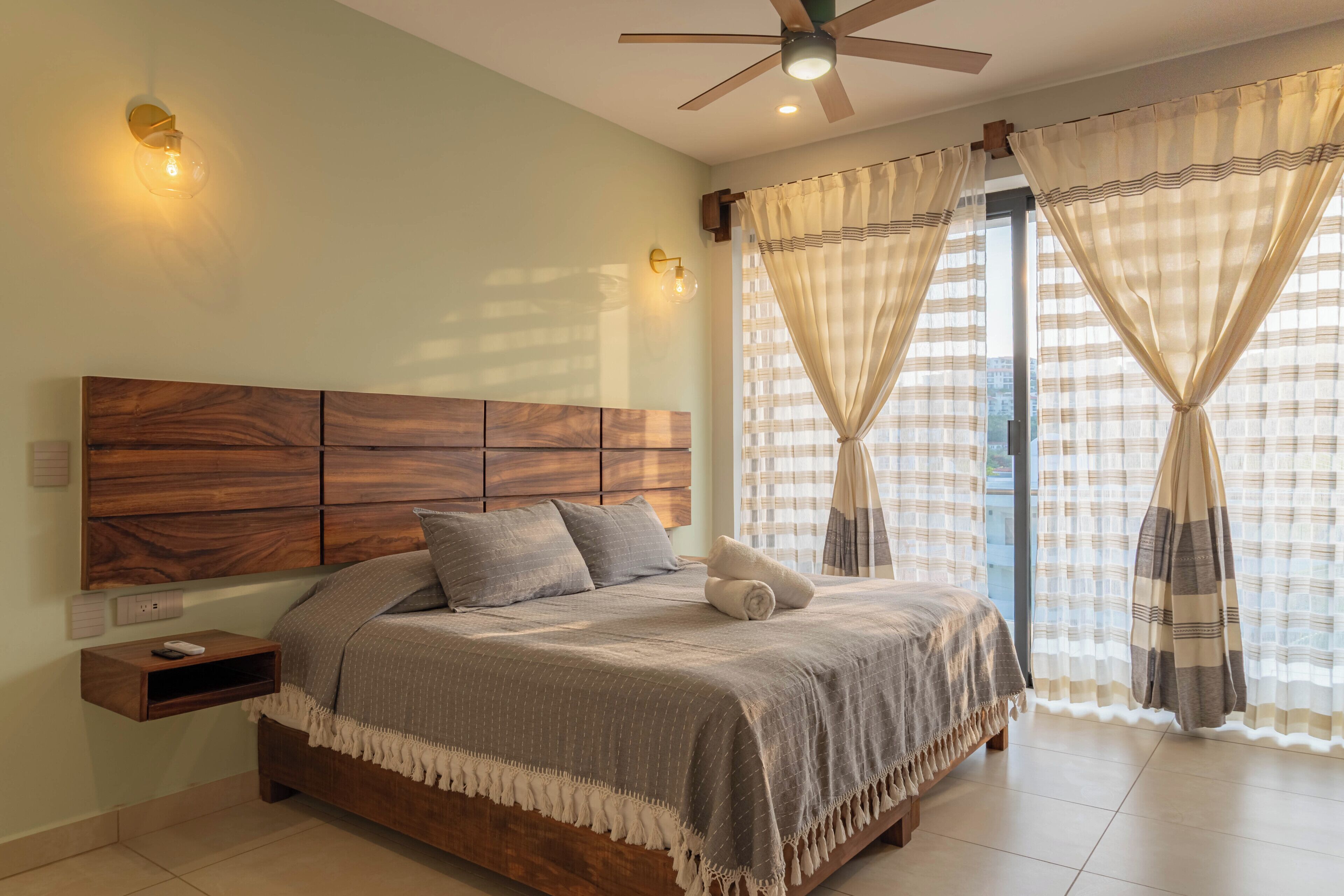 Una camera da letto con una testiera in legno, un letto con biancheria grigia, un comodino e una ventola a soffitto.