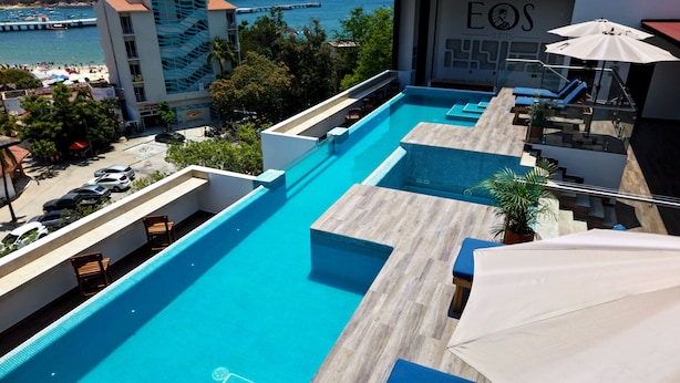 A rooftop pool with clear blue water, surrounded by lounge chairs and umbrellas, overlooking a beach and a pier. - EOS Suites (Santa Maria Huatulco)