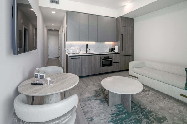 A modern kitchen with grey cabinets, a built-in oven, and a coffee machine. There is a round dining table with two chairs, a sofa, and a small rug on the floor.