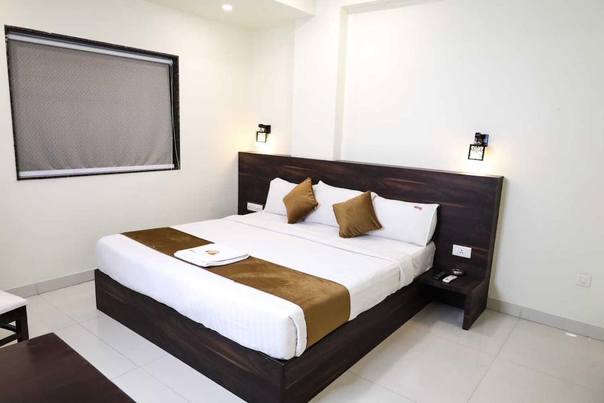 A neatly made bed with a brown headboard, white bedding, and a brown throw blanket. There are two bedside tables with lamps, a window with a blind, and a wall-mounted light fixture.