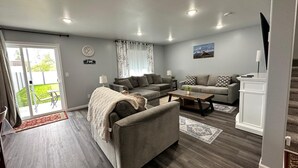 A modern living room with a grey sofa, a coffee table, and a TV stand. There is a sliding glass door leading to a patio with a table and chairs.