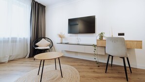 Ein modernes Wohnzimmer mit einem runden Holztisch, zwei Stühlen, einem an der Wand befestigten Fernseher und einer Pflanze auf der Kommode.