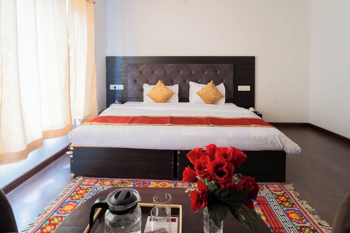 A neatly made bed with a dark headboard, a colorful rug, and a vase of red flowers on a wooden table.