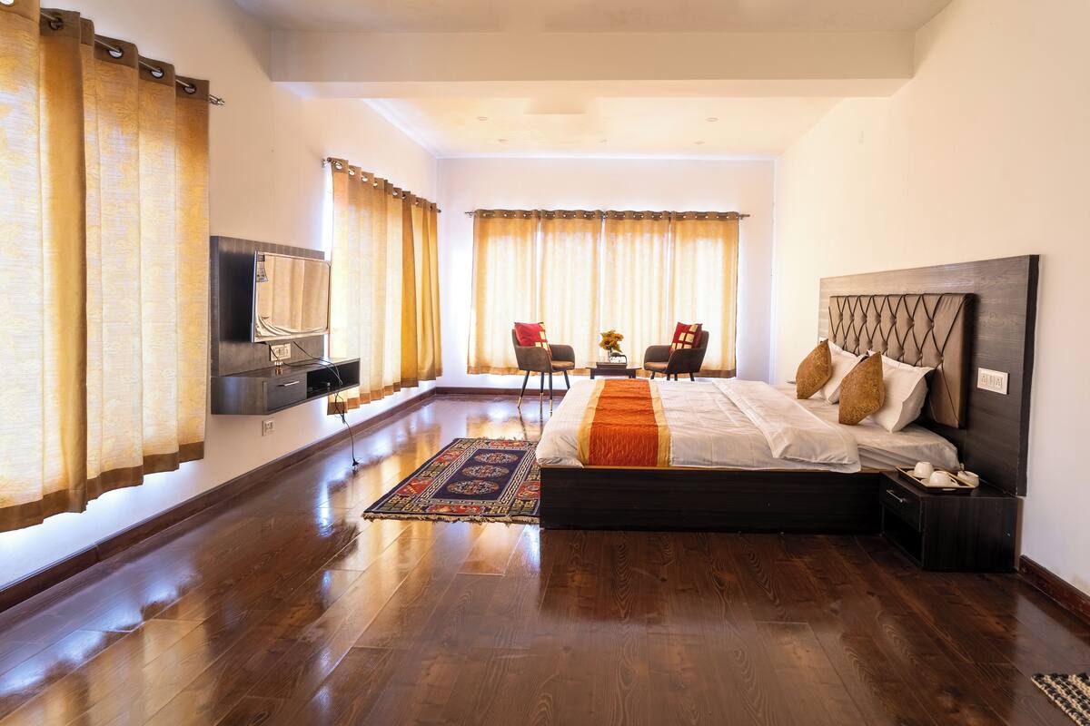 A spacious bedroom with a large bed, a TV mounted on the wall, a desk with two chairs, and a view of the outdoors through large windows.