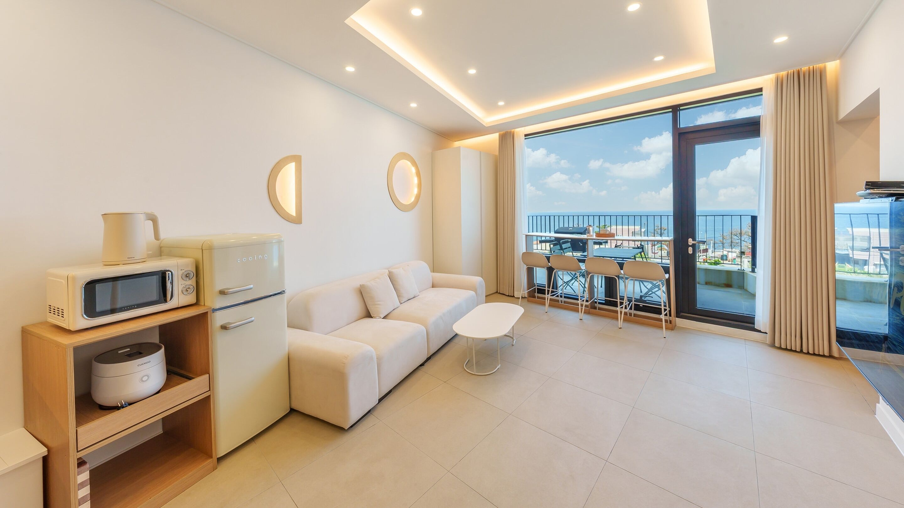 A modern living room with a sofa, a coffee table, a microwave, and a refrigerator. There is a balcony with a table and chairs.