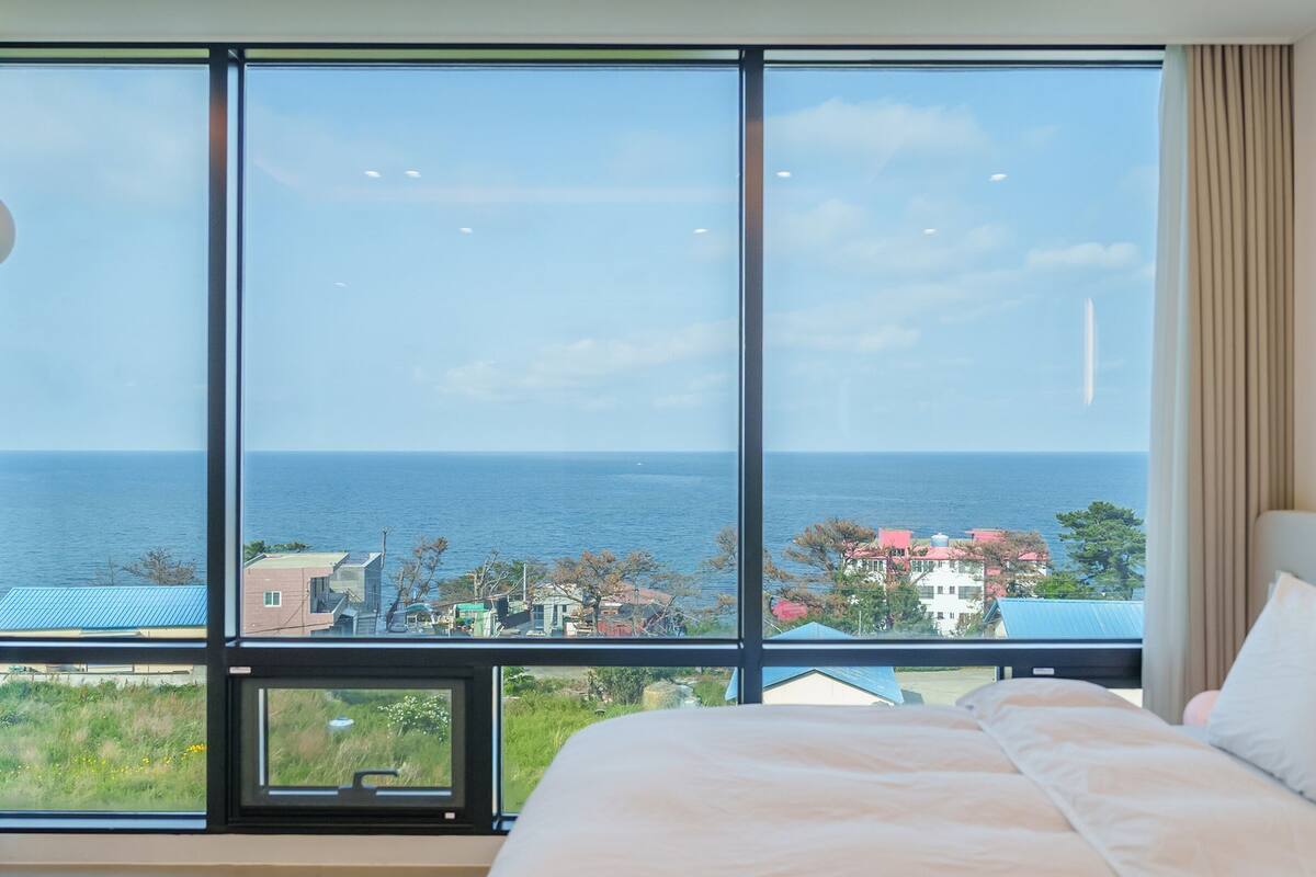 A bedroom with a large window offering a view of the sea and buildings.