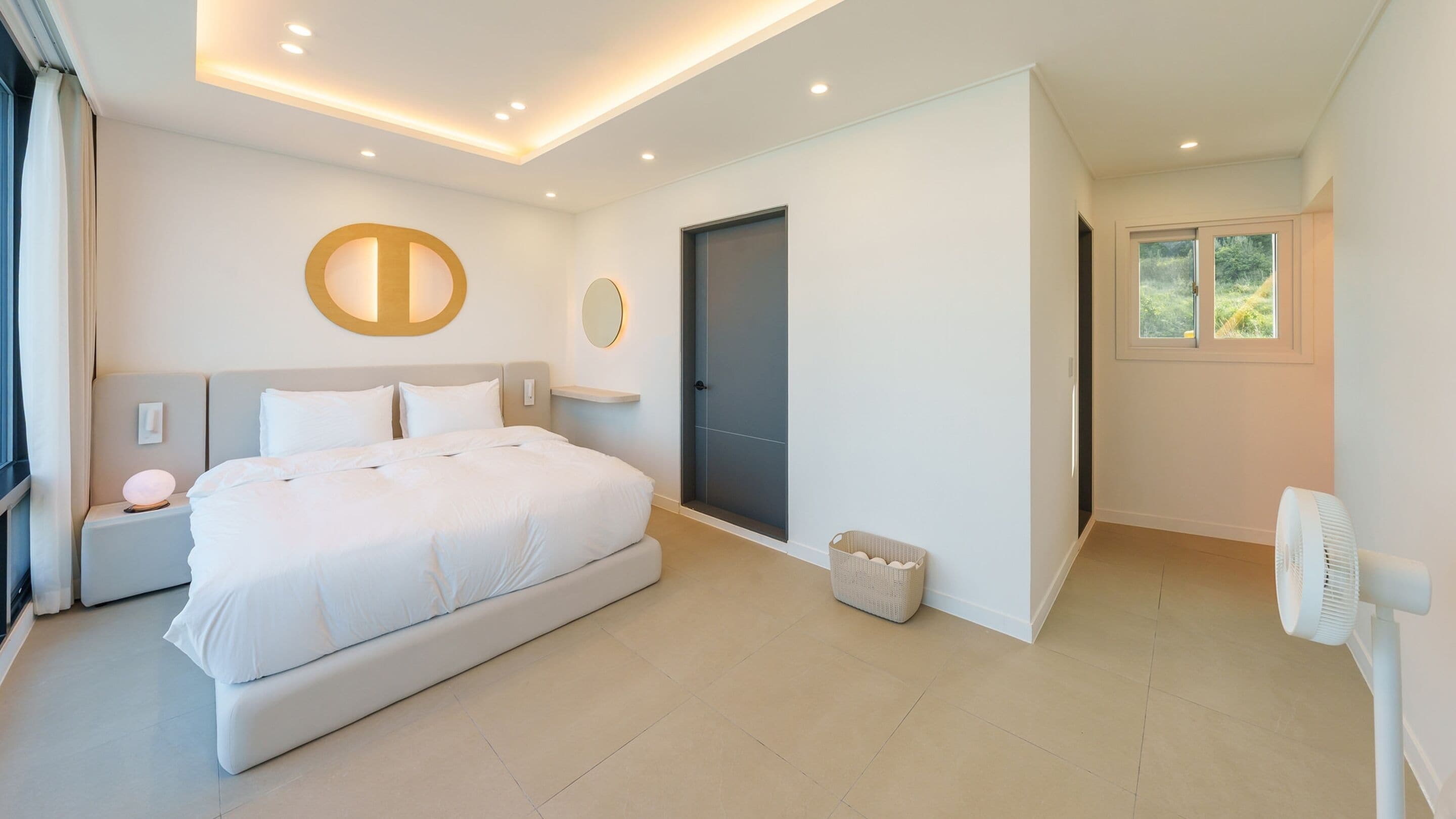 A modern bedroom with a large bed, a bedside table, a nightstand, a wall-mounted light, a door, and a window.