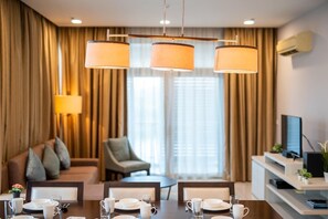 Ein Hotelzimmer mit Esstisch für vier Personen, einer Couch, einem Sessel, einem Fernseher und einer Klimaanlage.