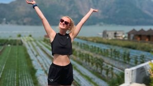 Une personne se tient sur une terrasse, les bras levés, et contemple un paysage composé de montagnes et d’une étendue d’eau.