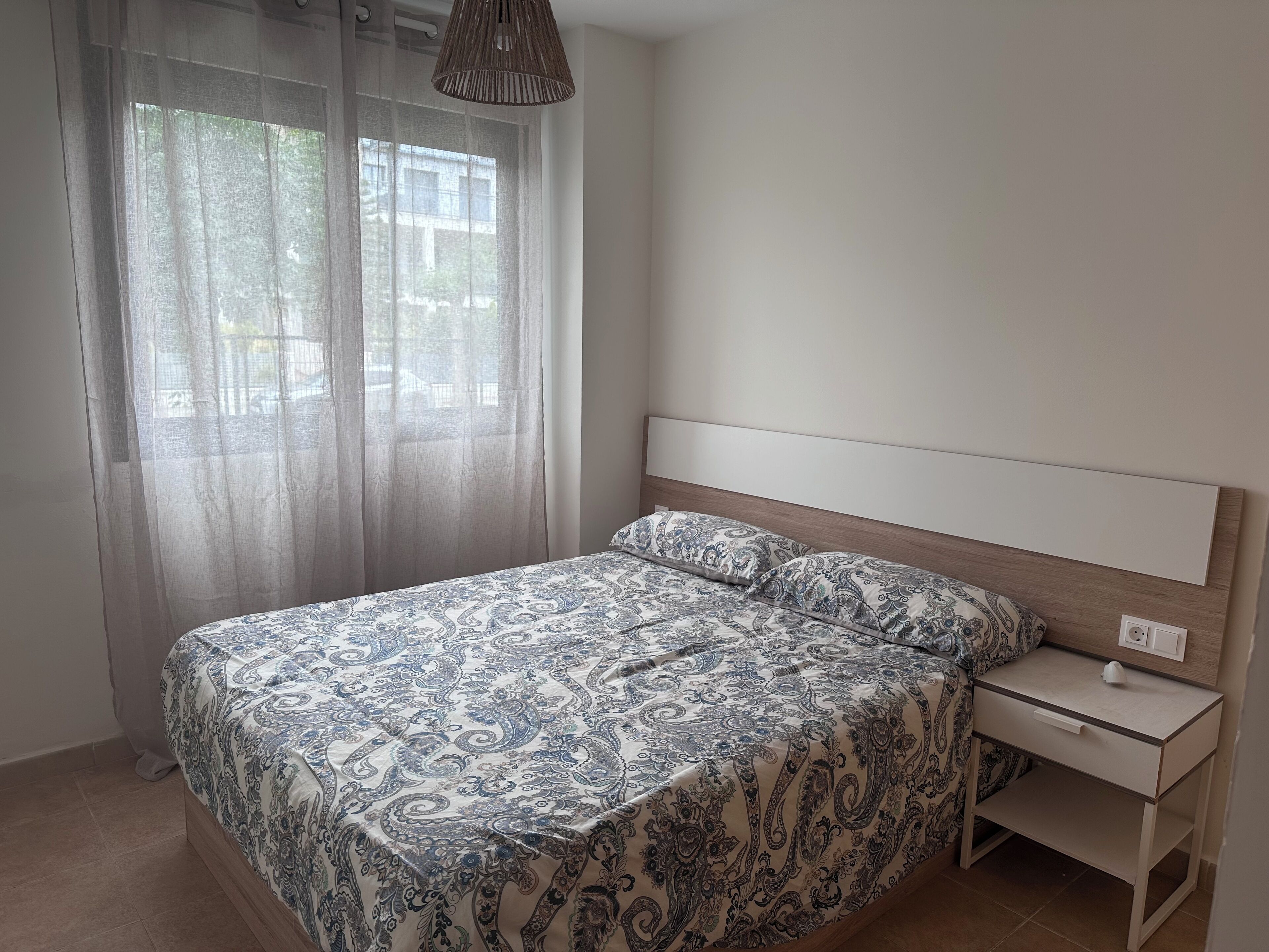 Un dormitorio con cama, mesita de noche, lámpara y ventana con cortinas translúcidas.