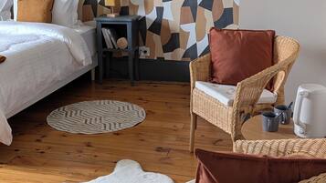 A bedroom with a geometric patterned wall, a bed with white bedding, a wicker chair with a brown cushion, a small table with a lamp, and a white rug.