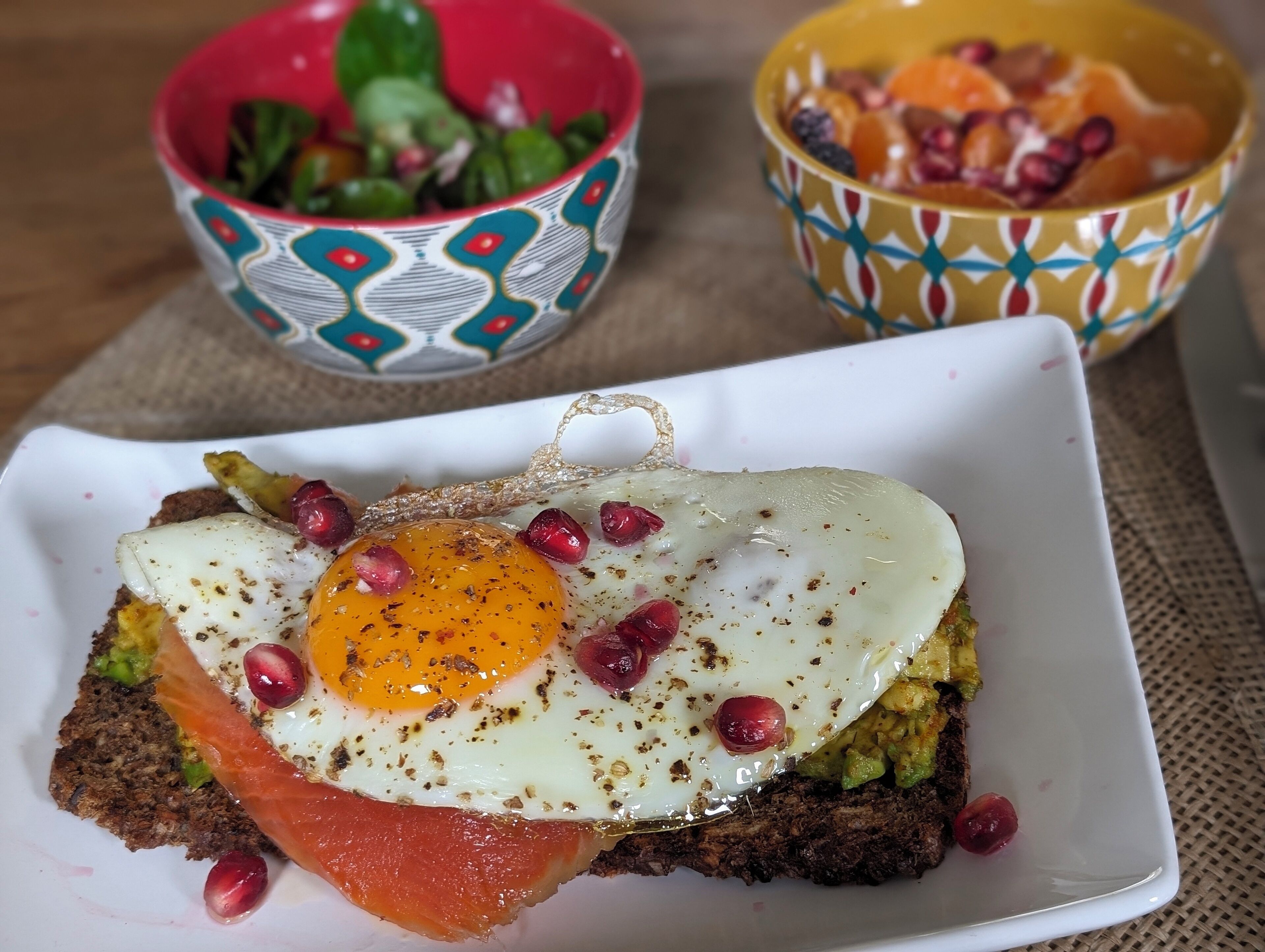 Une assiette de petit-déjeuner avec un œuf au plat, de l’avocat et du saumon fumé sur une tranche de pain, accompagnée de deux petits bols décoratifs remplis de fruits.