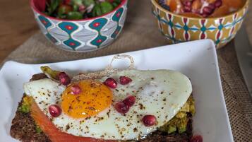 Une assiette de petit-déjeuner avec un œuf au plat, de l’avocat et du saumon fumé sur une tranche de pain, accompagnée de deux petits bols décoratifs remplis de fruits.