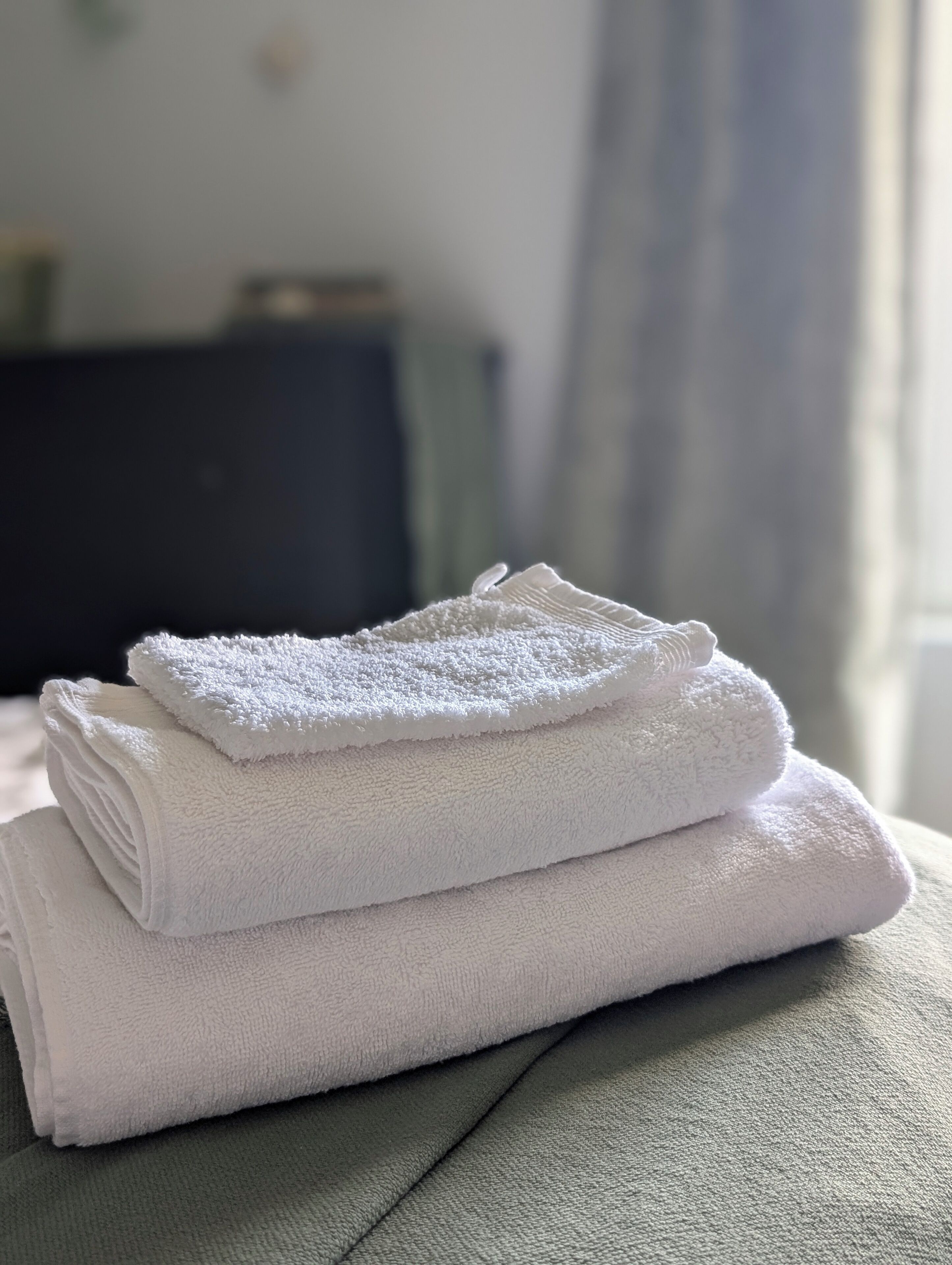A neatly folded white towel stacked on a textured grey surface, likely a bed or couch, in a room with a dark piece of furniture in the background.