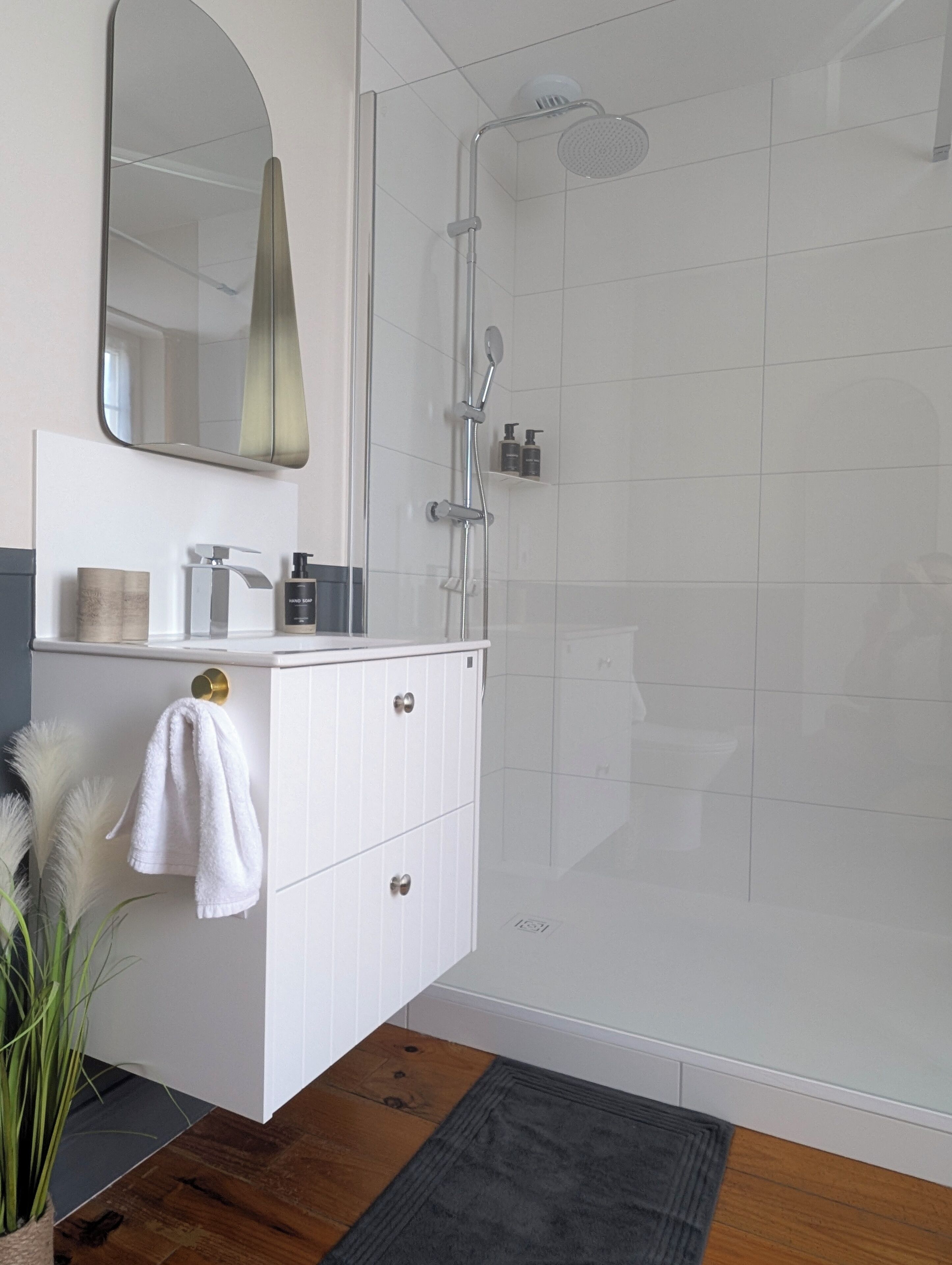 Un baño moderno con un tocador blanco, un espejo grande, una ducha con cabezal de ducha manual y un estante pequeño con artículos de aseo.