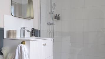 Un baño moderno con un tocador blanco, un espejo grande, una ducha con cabezal de ducha manual y un estante pequeño con artículos de aseo.