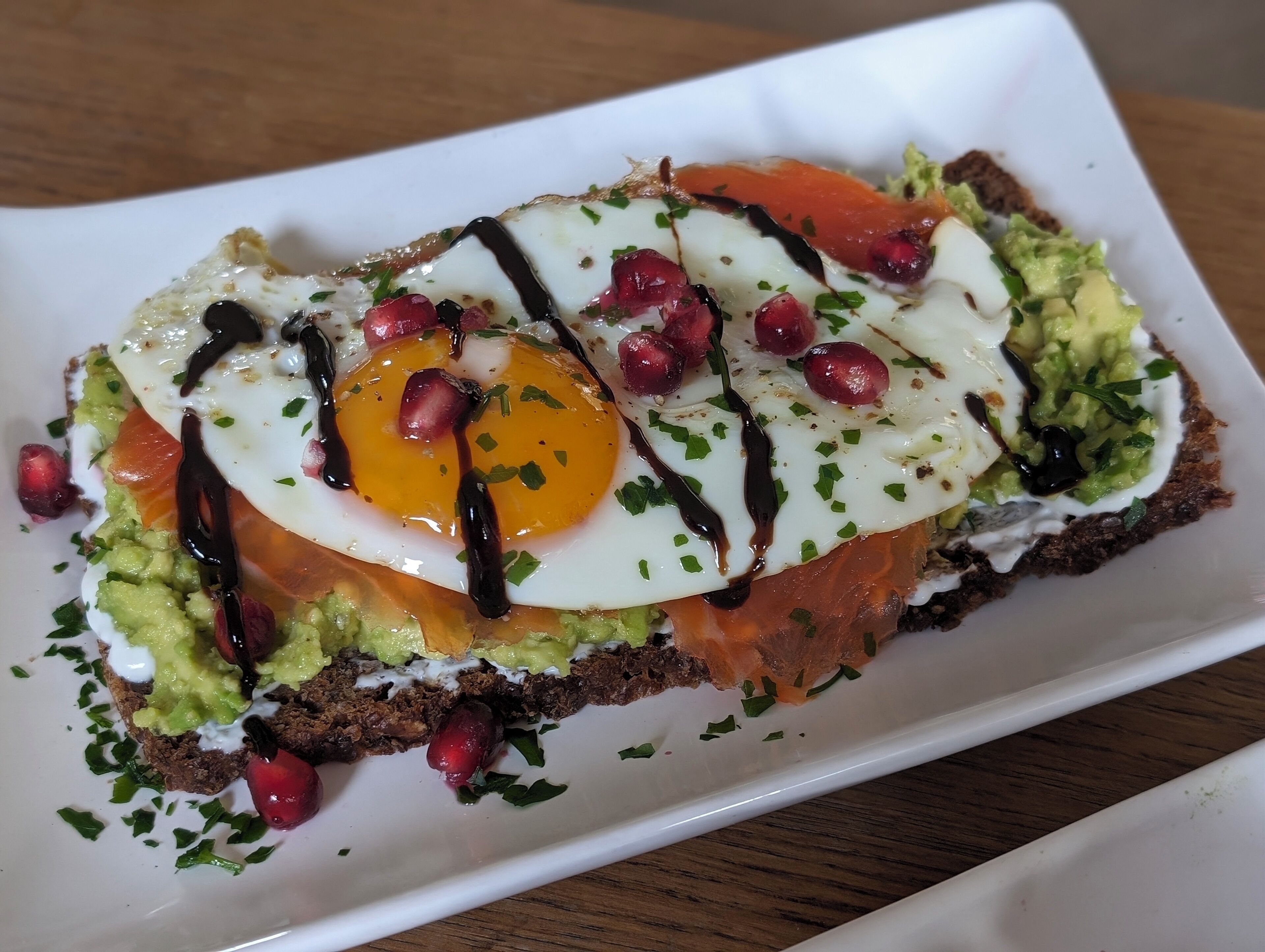 Un plato con aguacate, huevo, granada y un chorrito de salsa sobre una rebanada de pan.