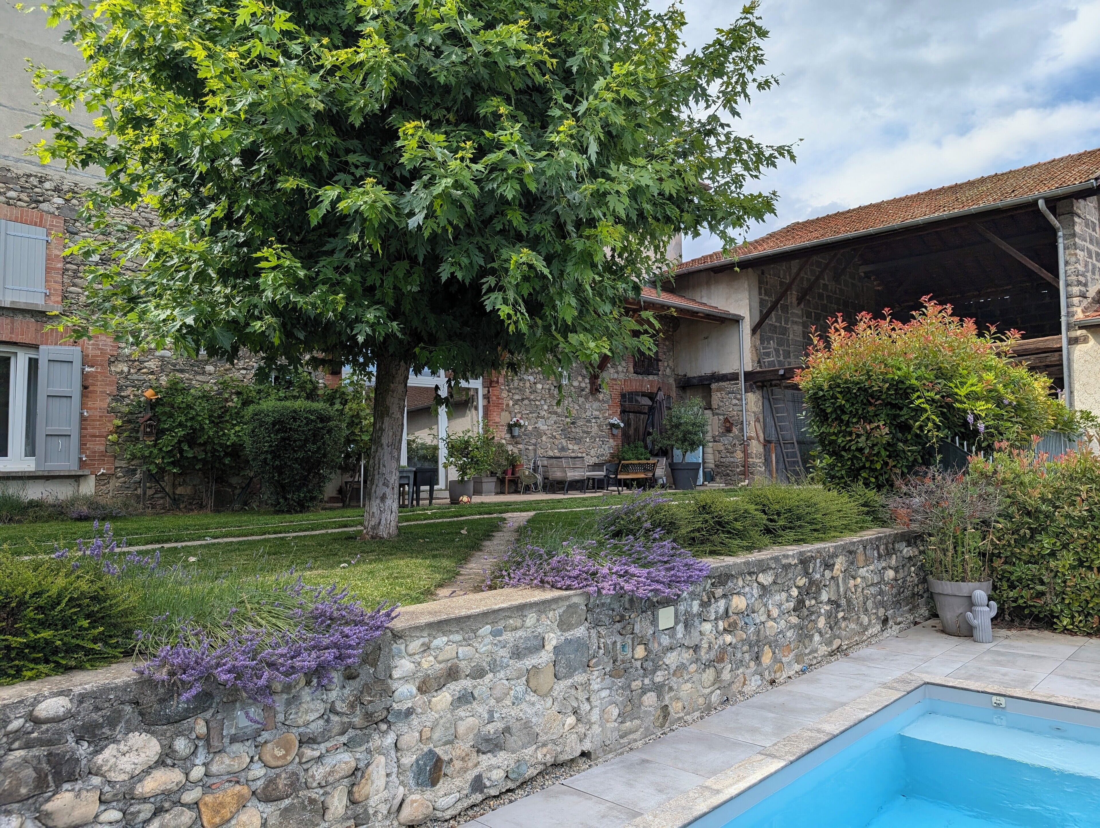 Un mur de pierre avec une piscine, un arbre et un bâtiment en pierre doté d’un toit en tuiles.
