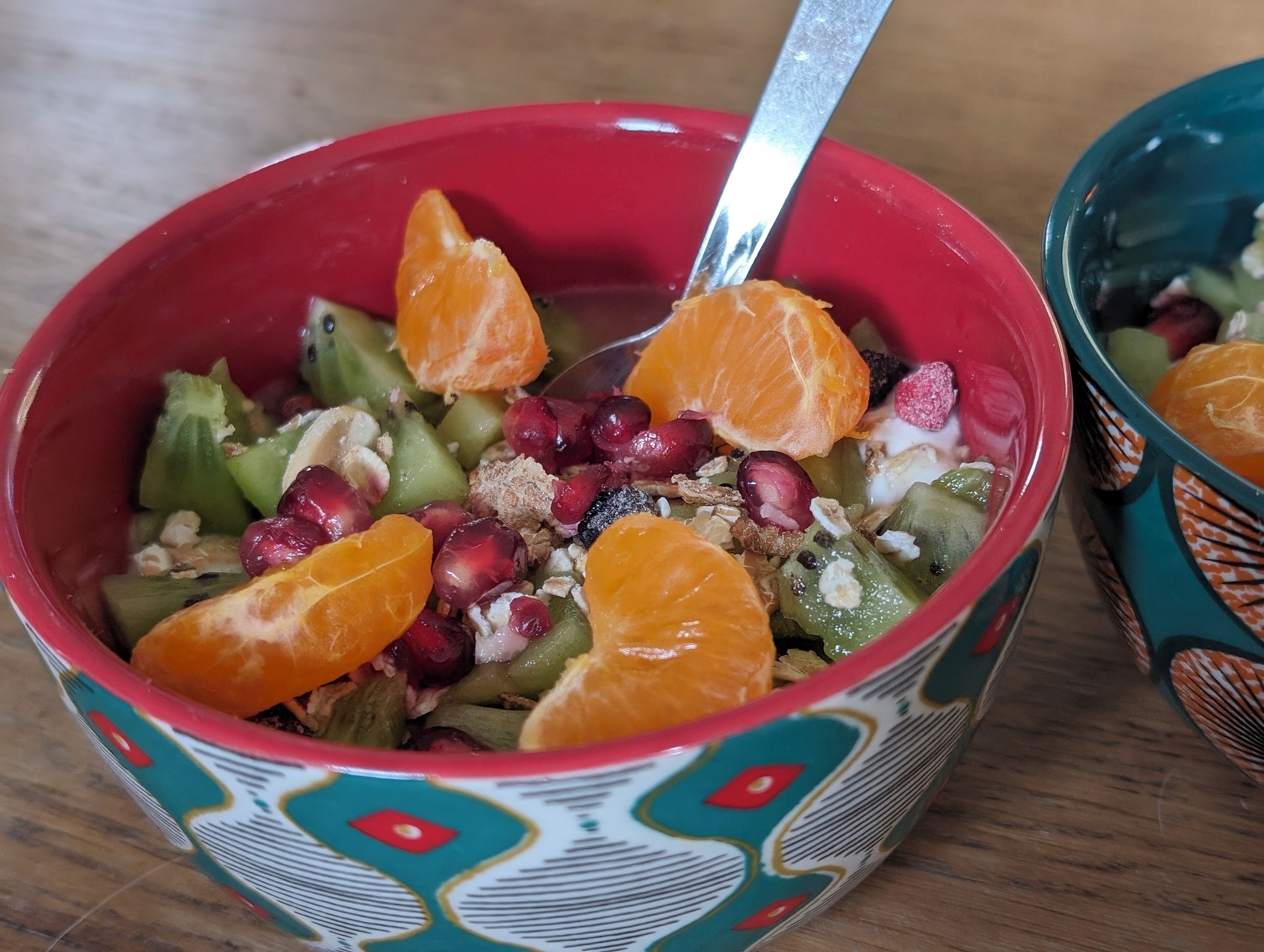 A bowl of fruit salad with a spoon, containing oranges, pomegranate seeds, and other fruits, in a colorful bowl with a geometric pattern.