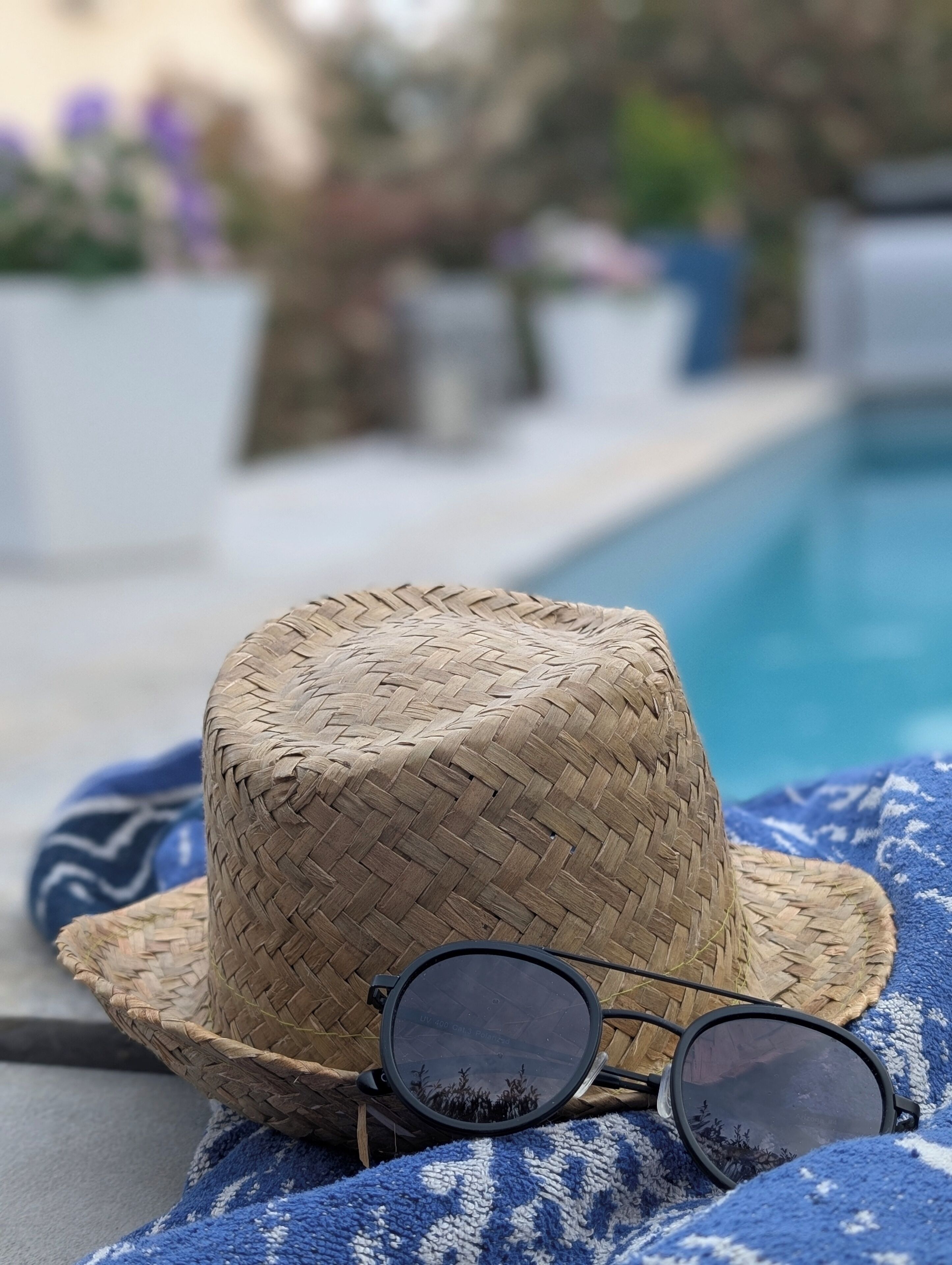A woven hat and sunglasses on a blue and white striped towel by a pool.