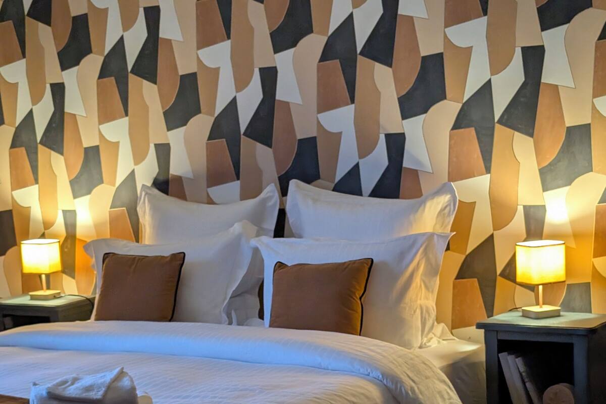 A neatly made bed with white linens and brown pillows, a round rug, and a geometric-patterned wall behind the bed.