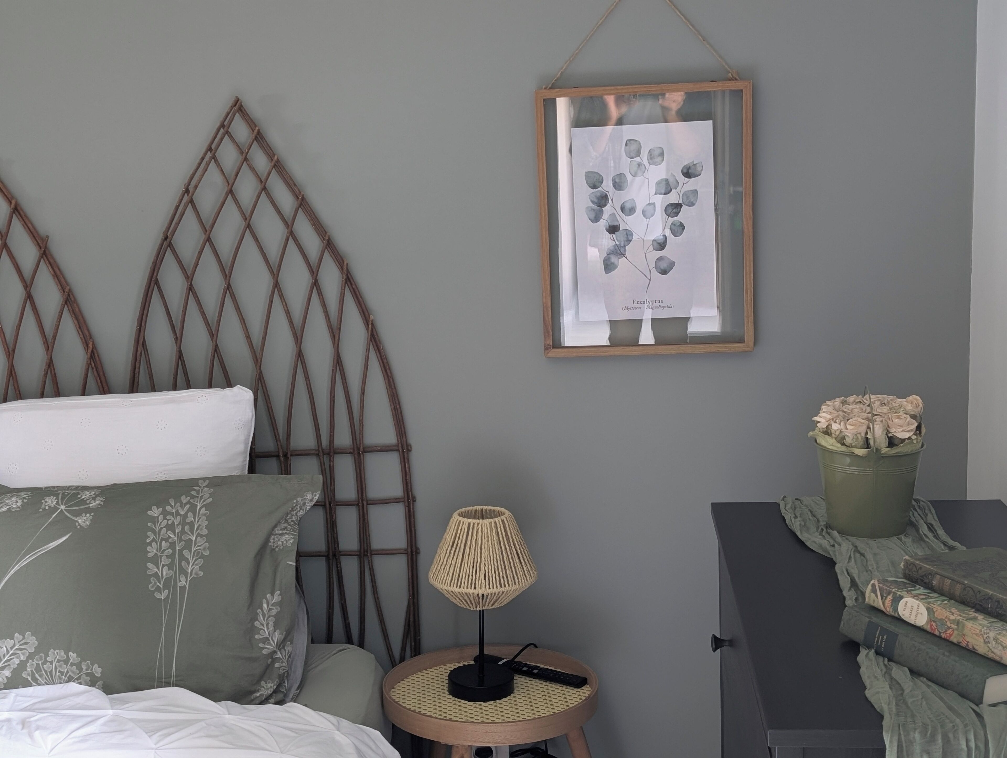 Une chambre à coucher avec un lit, une estampe botanique encadrée, une petite table avec une lampe et une commode sur laquelle est posée une plante en pot.