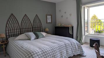 A bedroom with a checkered bedspread, a wooden headboard, a nightstand with a lamp, a dresser, and a window with a view of greenery.