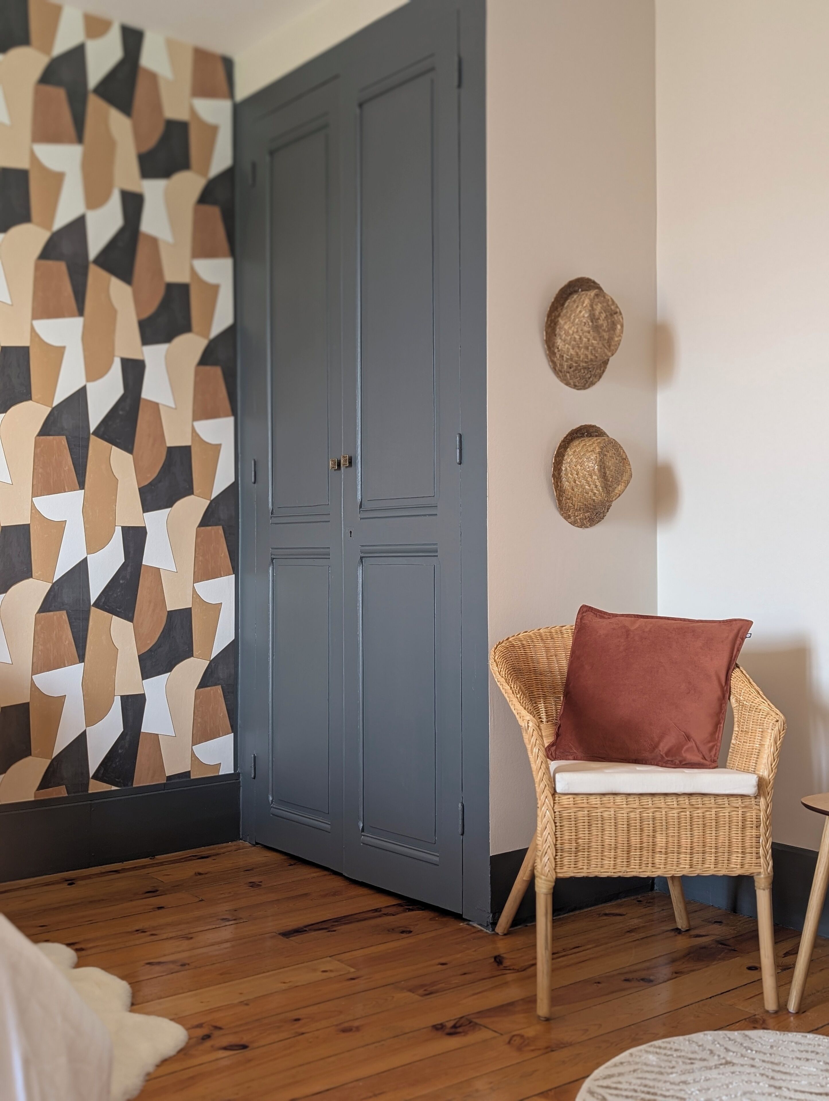 Een kamer met een wand met een geometrisch patroon, een grijze deur, een rieten stoel met kussen en een ronde tafel.