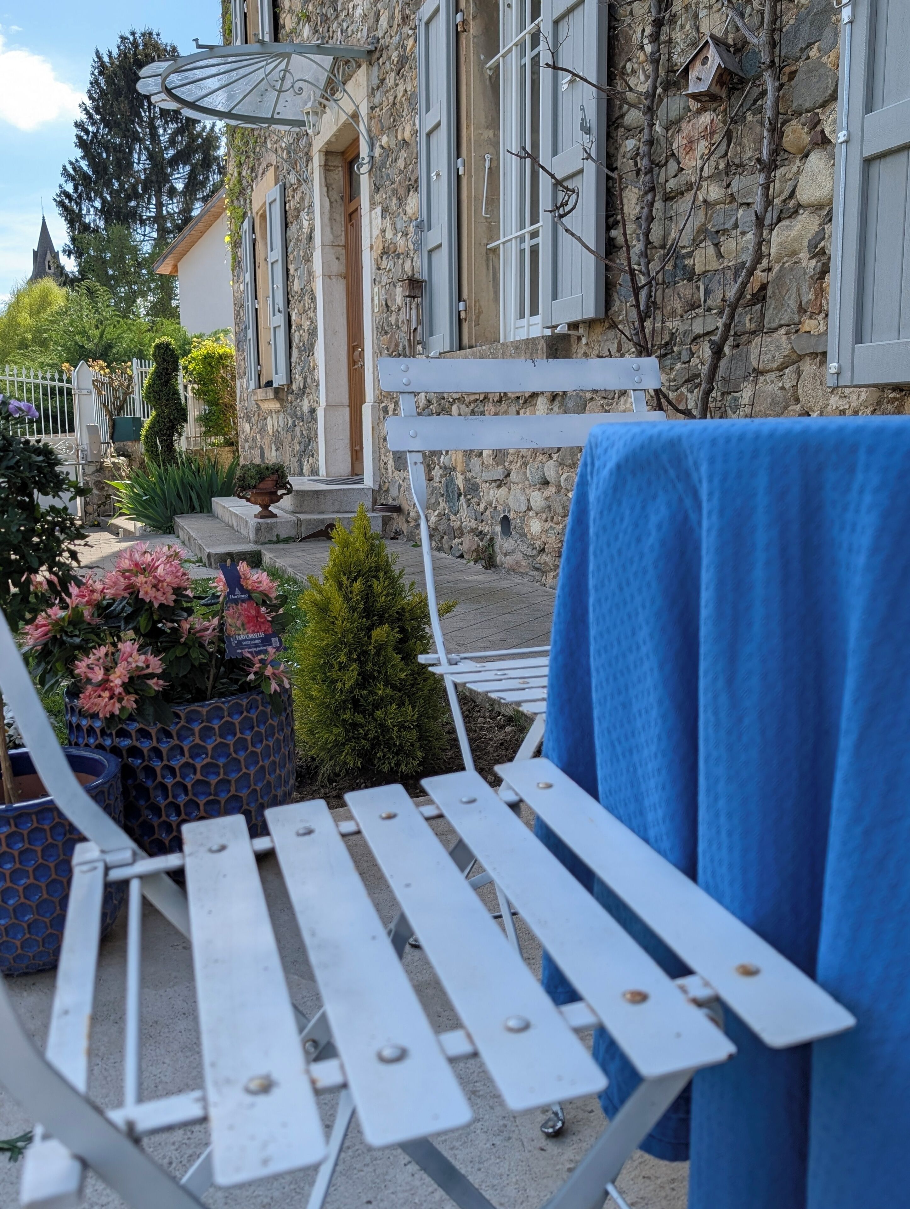 Un espace extérieur aménagé avec des sièges et une table recouverte d’une nappe bleue, entouré de plantes en pot et d’un bâtiment en pierre doté d’une porte en bois.