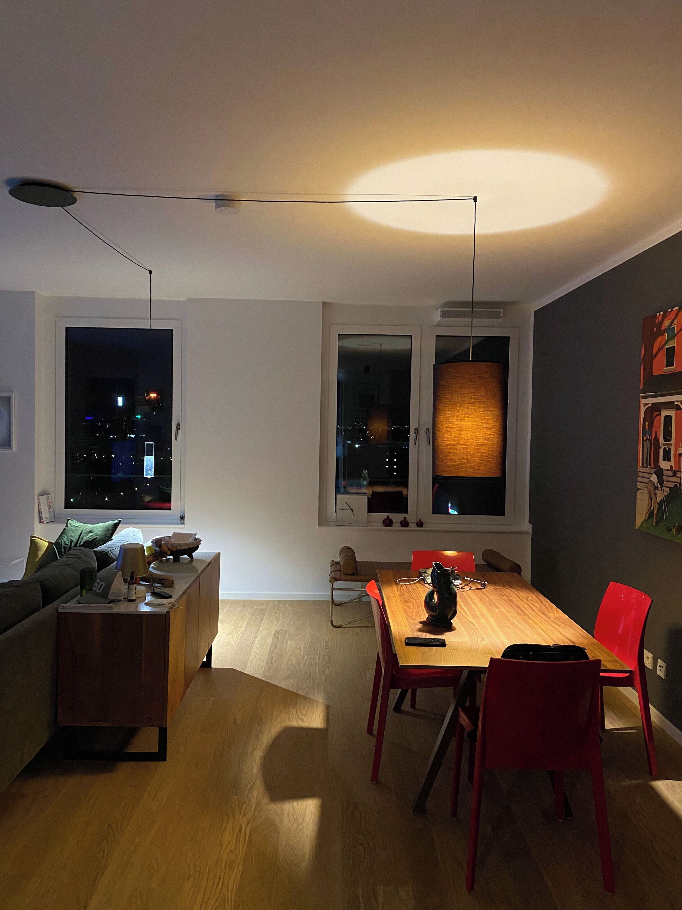 A modern living room with a dining area, featuring a wooden dining table, red chairs, a sofa, and a large window with a city view at night.
