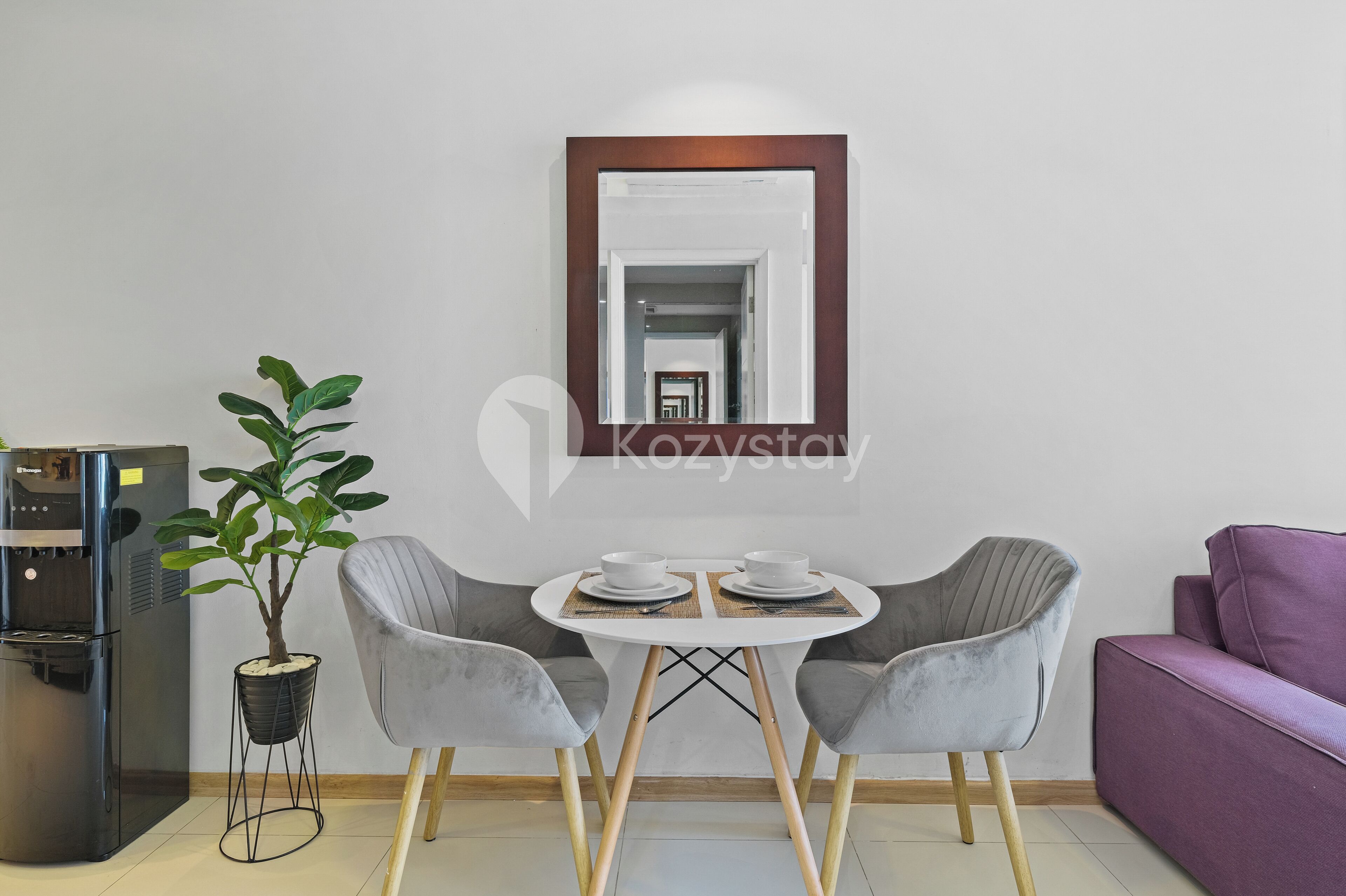 A modern living room with a white table, two grey chairs, a purple sofa, a coffee machine, and a potted plant.
