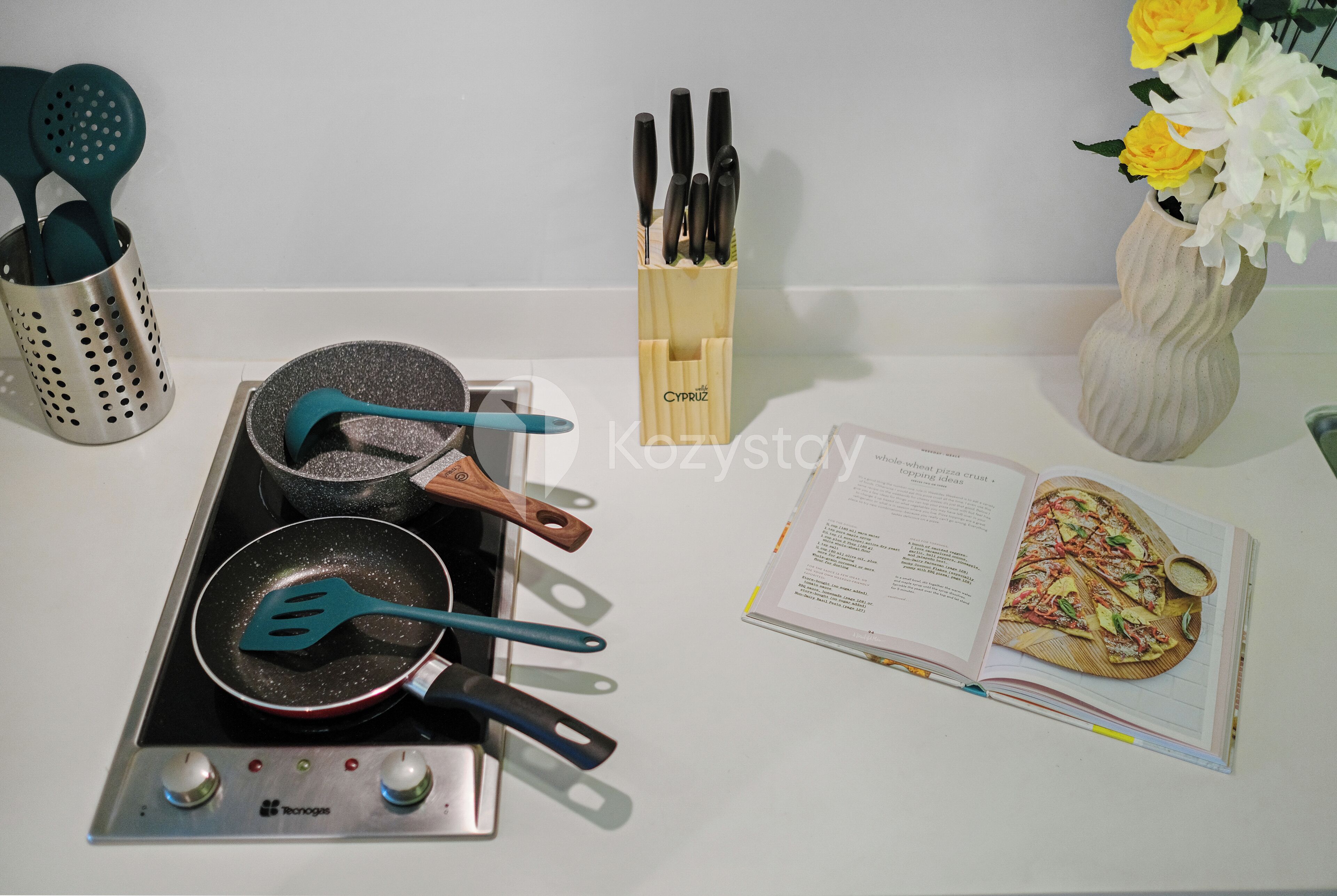 Una encimera de cocina con estufa, sartenes, espátulas, un porta cuchillos y un libro de cocina.