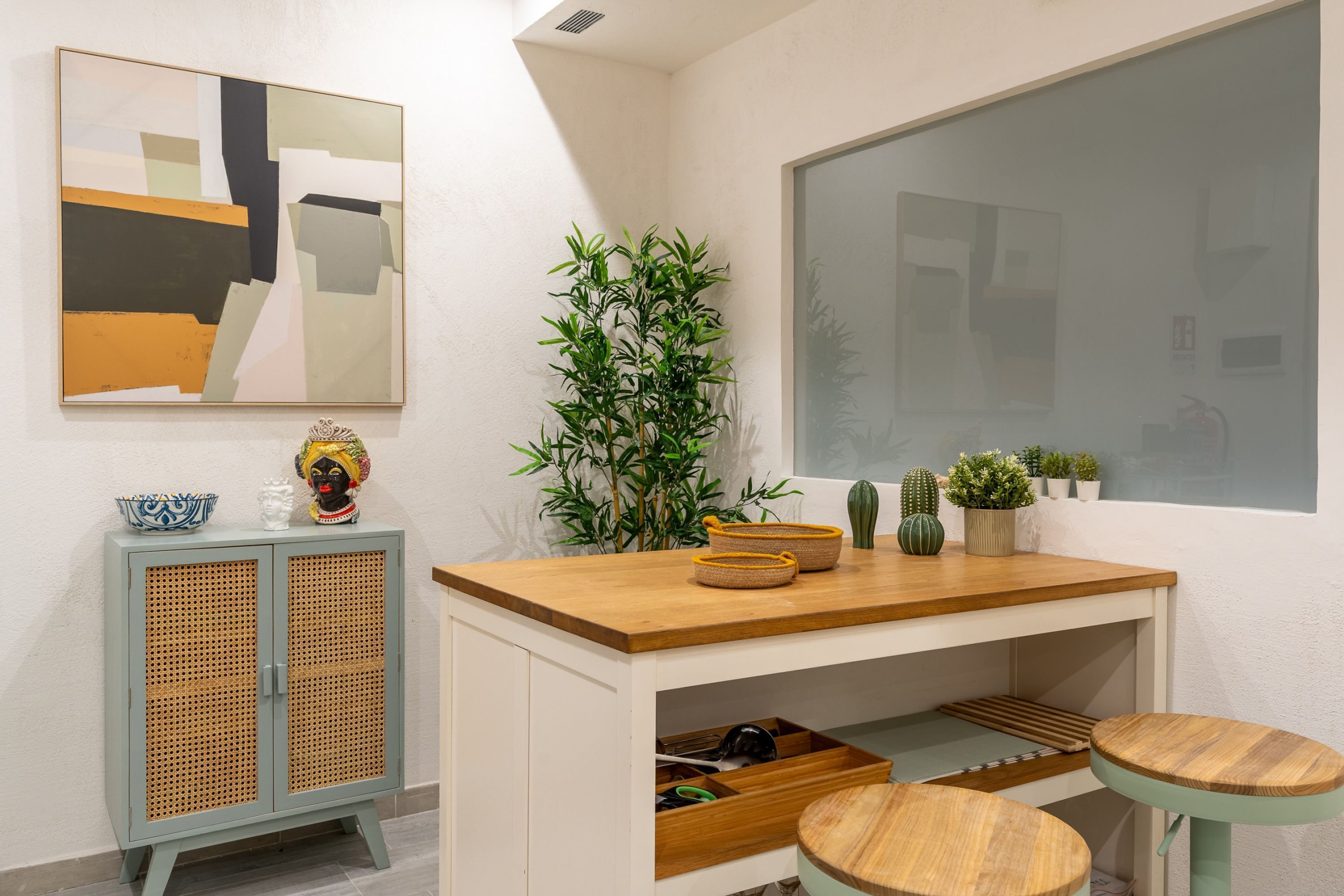 Une pièce moderne avec un bureau en bois, deux tabourets, un meuble en rotin et un grand tableau abstrait accroché au mur.