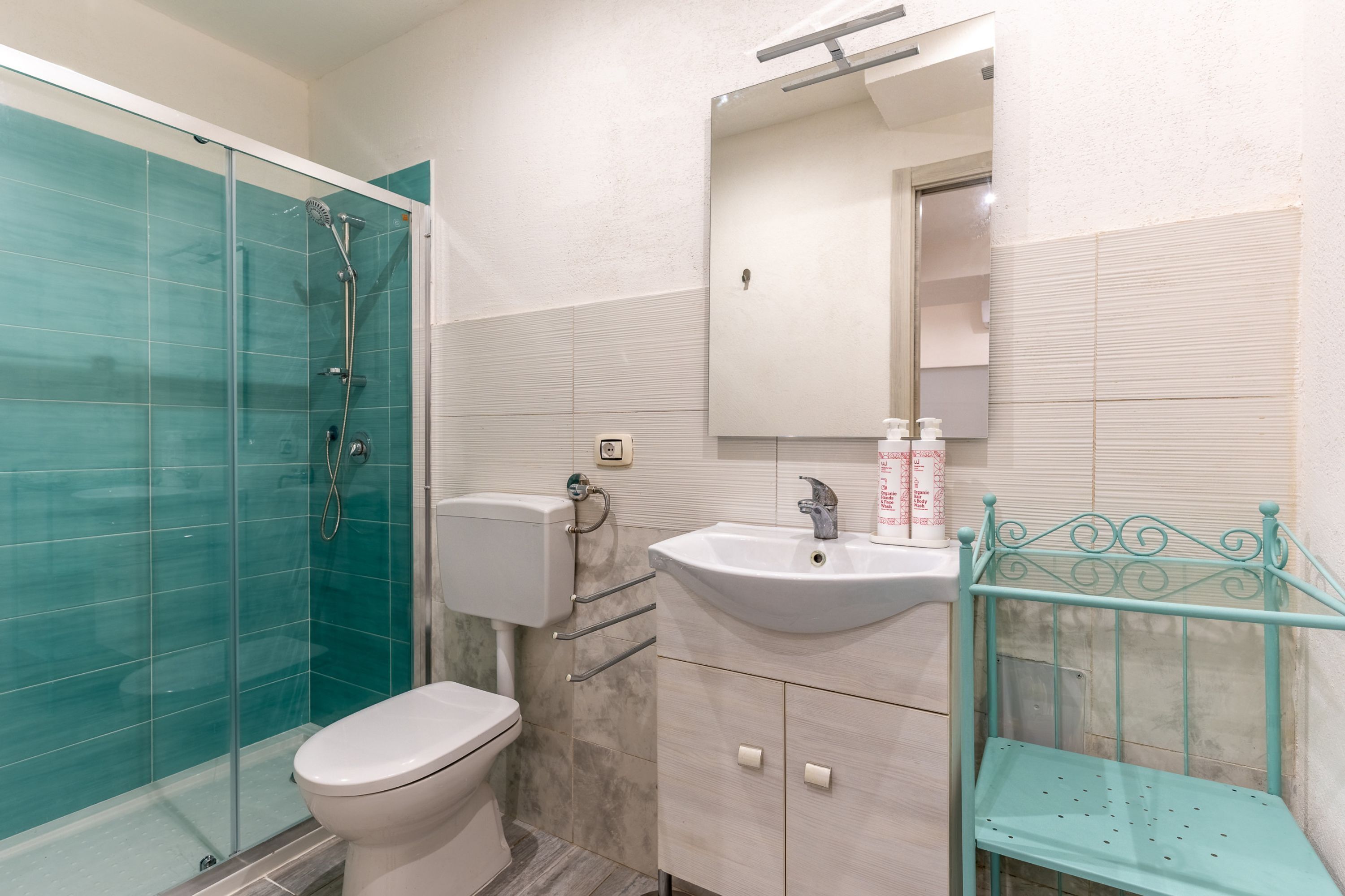 A bathroom with a glass shower enclosure, a toilet, a sink with a mirror above it, and a metal shelf.