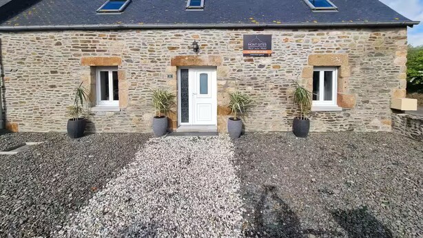 Il s’agit d’un bâtiment en pierre, doté d’un toit en pente, de trois fenêtres et d’une porte. Devant chaque fenêtre, on trouve des plantes en pot.