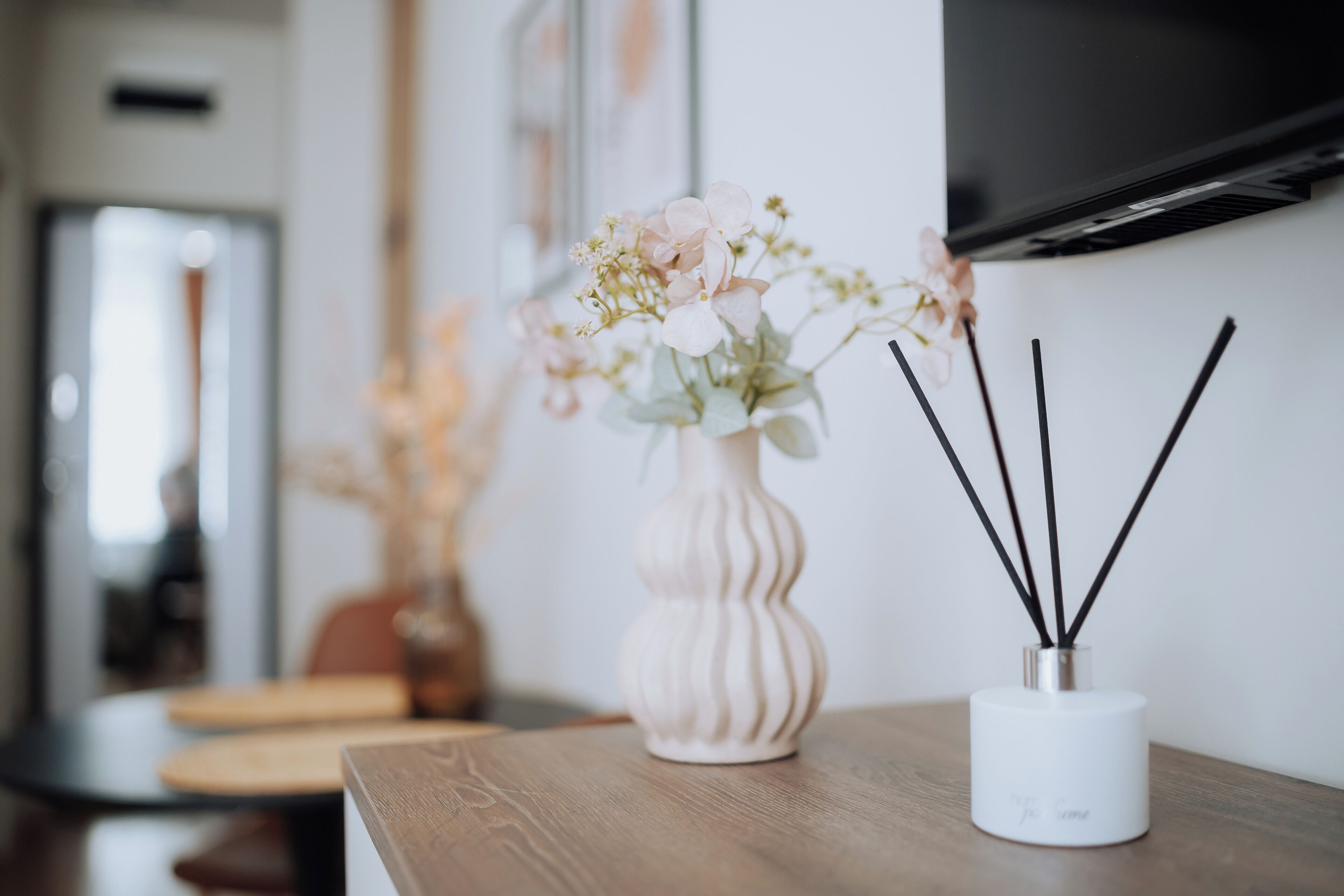 A modern living room with a wooden table, a white vase with flowers, and a white diffuser with reed sticks.