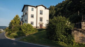 A two-story building with a central entrance, surrounded by greenery and a road.