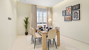 A modern dining room with a wooden table, beige chairs, a potted plant, and a large window with sheer curtains. - Silkhaus Osus 7 (Riyadh)