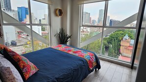 A bedroom with a large bed, a view of the city through floor-to-ceiling windows, and a wooden floor. - A 3 cdras Reforma/Angel/Embajada USA con Elevador (Ciudad de México)