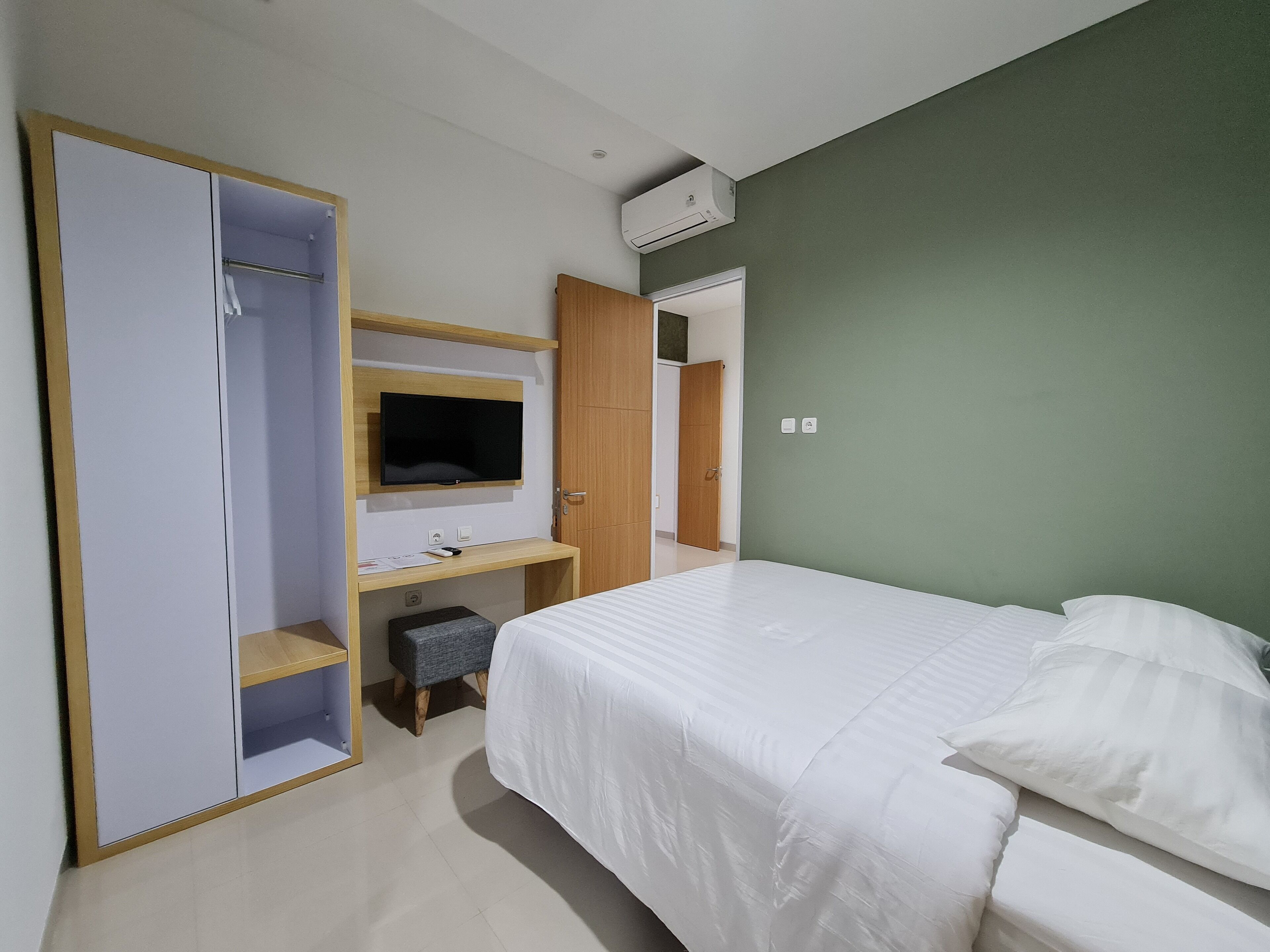 A bedroom with a bed, a TV mounted on a wooden shelf, a desk with a chair, and a wardrobe.