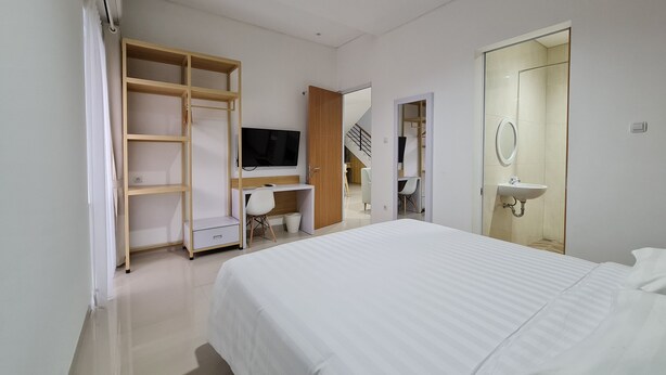 A modern bedroom with a bed, a desk with a chair, a TV, a wardrobe, and a bathroom with a sink and mirror. - Roemah Audy (Pogung Lor)
