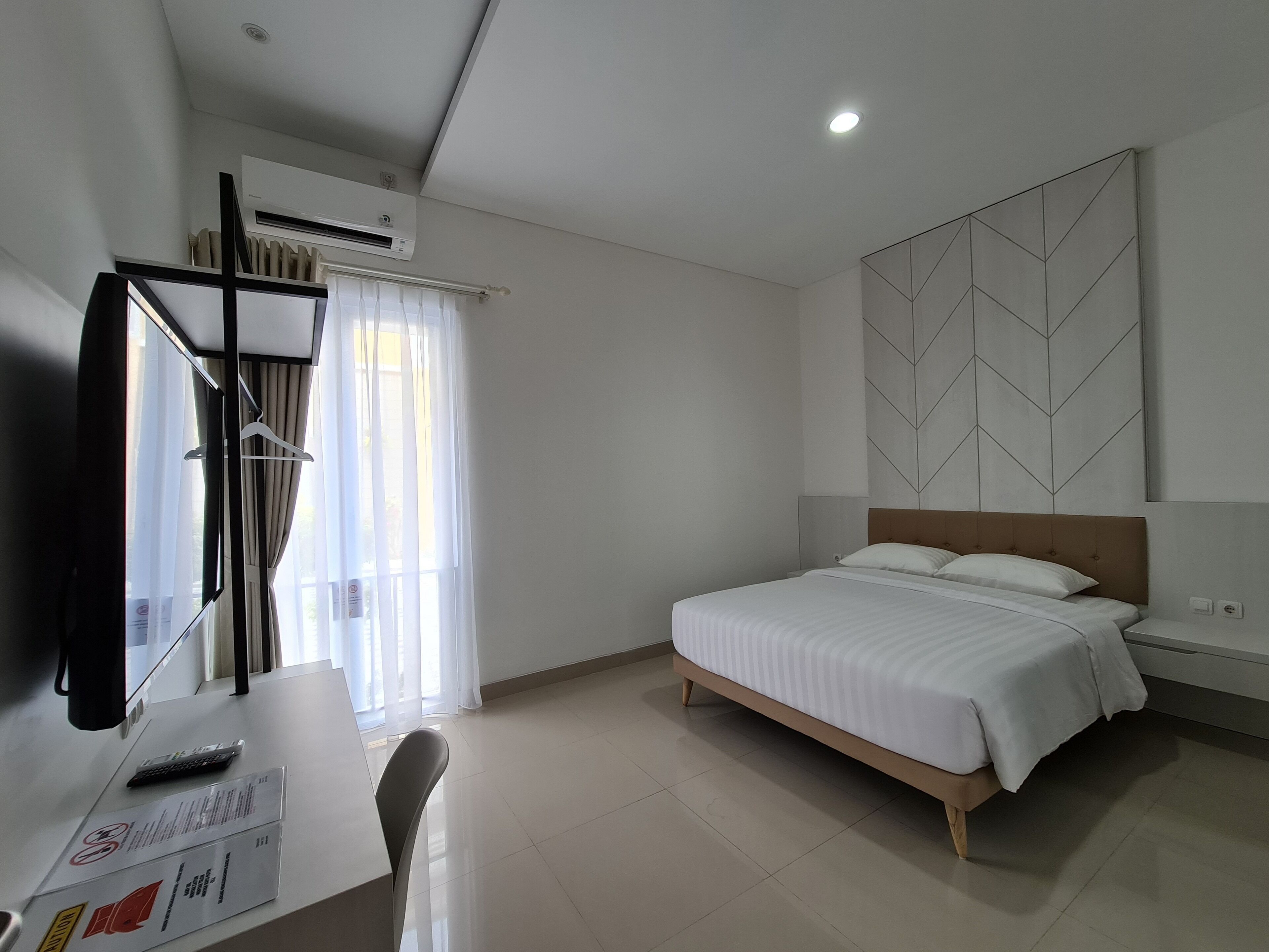 A hotel room with a bed, a desk with a computer, an air conditioner, and a window with curtains.