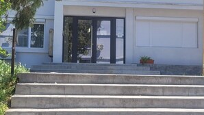 Ein Gebäude mit einer Treppe aus Beton, die zu einem Doppeltür-Eingang führt. Auf den Stufen steht ein kleiner Blumentopf und links daneben ein Baum.